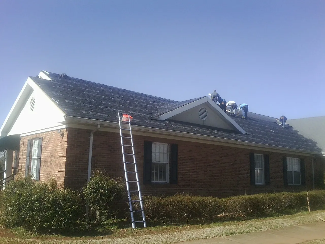 Crew tearing shingles off a brick ranch roof with a ladder set — Emergency Roof Repair work by our crew in Rienzi