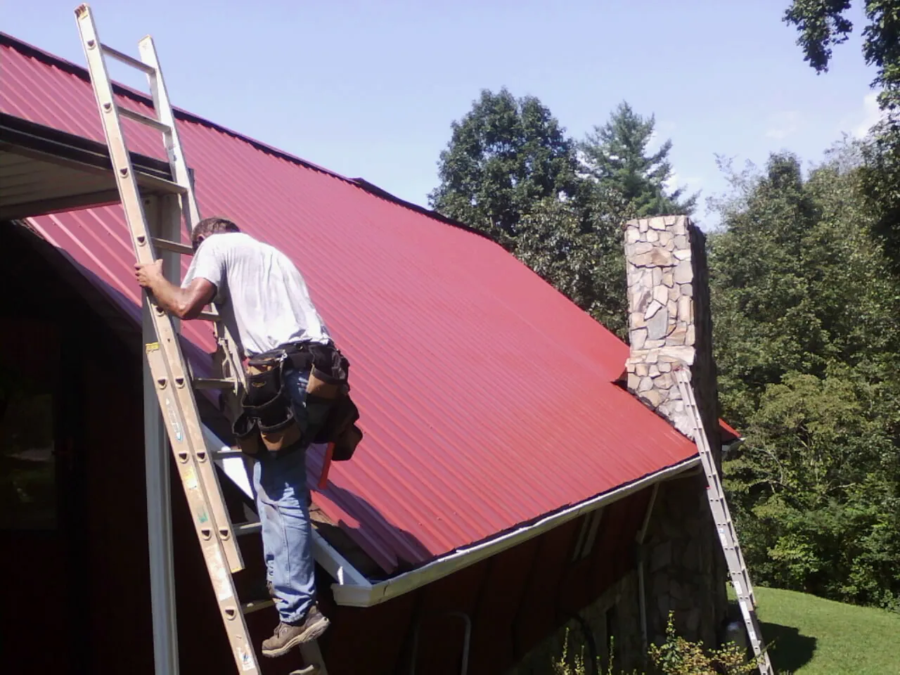 Roofer on a ladder beside a red metal roof and stone chimney — Soffit & Fascia Repair by Roofing Brothers in Rienzi